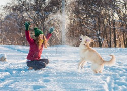 Happy beautiful young woman blowing snowflakes from her hands to her dog golden retriever in a winter day. Friendship, pet and human.