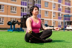 young sturdy chubby woman meditates on the lawn after exercising in the courtyard of a city house against the background of street gym equipment