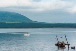 waterscape of the sea bay with a sailing ship and shipwreck and a mount in the clouds in the background, a view of the Mendeleev volcano from the side of the town of Yuzhno-Kurilsk