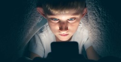 Boy with a serious face under the blanket at night in his bed communicates on Internet. Child gadget addiction and insomnia.