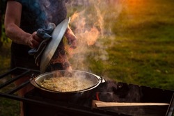 A woman cook opens the lid of a saucepan or cauldron in which pilaf is cooked on an open fire in the grill. Steam rises from the cauldron. Natural grass background.