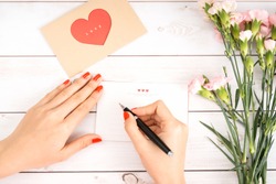 Woman writes love letter on white paper with red heart shape figures.Hand made postcard for Saint Valentines Day celebration.Send letters for your lovers & friends on 14th of February