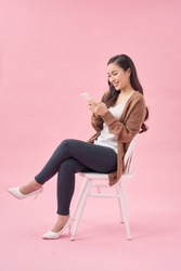 Portrait of a satisfied casual asian woman using mobile phone while sitting on a chair over pink background