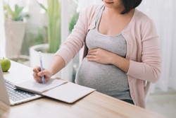 Closeup of a pregnant woman writing notes and using a laptop while working on maternity leave at her dining room table at home