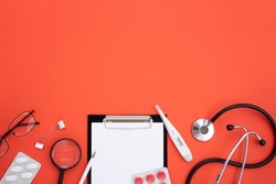 Top view Medic work desk with copy space. Tablet for notes, stethoscope, thermometer, pills, medicines, magnifier, glasses, on a red background. Flat lay, mockup