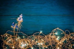 Glowing Christmas lights, silver beads and decorations deer and snowman on sticks on blue wooden background. Top view with copy space
