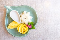 Thai dessert - Mango sticky rice and coconut milk sauce serving on lotus leaf shape plate, decorated with leaves and pink flowers, on beige fabric tablemat background, top view, horizontal, copy space