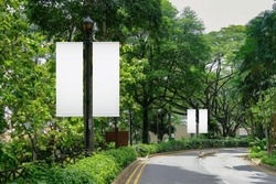 Blank vertical advertising banners on street lampposts; double hanging posters by the road, against lush green trees and plants. For OOH out of home template mock up.