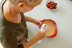 High angle view of mature caucasian woman mixing batter in bowl with electric whisk on kitchen island at home