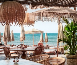 Beach club concept, food corner or cafe, restaurant tables, straw natural chairs, wooden tables, makrome knitted sun umbrellas, white modern sun beds, sea view on background