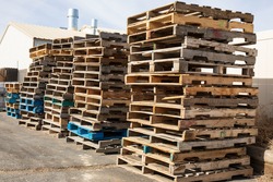 Wooden pallets stacked along a wall in a warehouse lot.  Stacks of pallets under a pale sky.  Loading dock with pallets piled high.