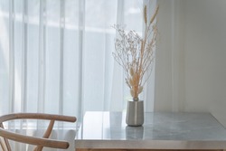 Beautiful dry flowers in an aluminum metal vase with light shines through the translucent curtains of the windows for decoration home interior on a wooden table. Concept modern building contemporary.