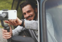 Portrait of successful man recording video at phone with steadycam. Smiling man sitting at the passenger seat of black car holding steadycam while recording steaming video sharing traveling experience