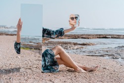 illusion portrait Stylish girl in sun glasses and dress sit on sea beach holding mirrors with reflection of sand and stones. New vision of fashion projects concept. Digital narcissism