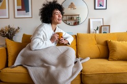 Happy African american woman enjoying quiet time at home laughing, drinking morning coffee sitting on sofa. Copy space.