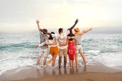 Rear view of multiracial friends embracing together looking at the ocean celebrating with arms up during vacation trip.