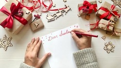 Woman's hands writing Christmas letter to Santa on white paper on wooden background with winter holiday decorations. Top view.