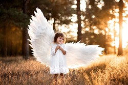 Cute little angel girl in white clothes, dress and wings, standing where the light of sunset, the sun's rays in the summer forest. Gentle, artistic image, photo, glare.