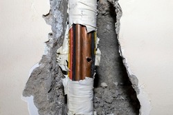 Damaged copper pipe in a wall surrounded by various wires and pipes