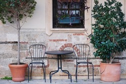 An elegance metal table and empty black chairs next to potted trees  in a street cafe  against the background of a window and a wall of an old stone building 
