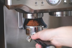 Hand holds a metal filter holder with grinded coffee beans in a coffee machine close-up, making espresso coffee