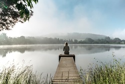 Calm misty morning relaxation by pond.Sitting woman in countryside.Intended female mysterious atmosphere. Spring foggy nature.Silence.Woman feeling freedom,enjoying vacation.No stress,calm mind,relax