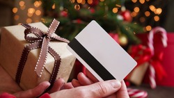 Young woman holding a credit card and a gift box against the background of Christmas decor and gifts, close-up. Christmas and New Year shopping on the Internet, payment by credit card.