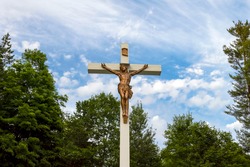 Big white wooden crucifix of Jesus Christ outdoors in a cemetery - bronze sculpture hit by sunlight against a blue sky with white clouds and green trees