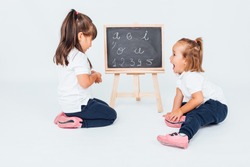Two girls, one with black hair and the other with blonde hair, in class, next to a chalk board, sniffing and playing