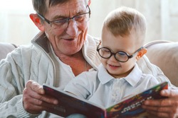 Elderly grandfather and little grandson read a book together and have fun sitting in a chair