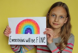 A girl on yellow background holds a sheet of paper. She drew a rainbow and wrote phrase Everything will be fine. The concept of the hope that the pandemic of coronavirus Covid-19 will finish soon