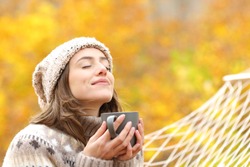 Relaxed woman breaths fresh air holding coffee cup for breakfast sitting on a rope hammock in autumn in a forest