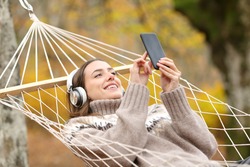 Happy woman with headphones and smart phone listening to music lying on hammock in autumn holiday