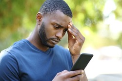Worried man with black skin checking smart phone standing in a park