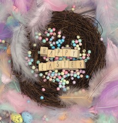 Colorful Easter eggs in bird nest surrounded with pastel colored feathers and wooden cubes with the text Happy Easter top view, modern background design, Easter holliday, Spring, concept greeting card