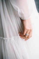 Right hand of the bride with engagement ring on her finger with her classy dress on background.