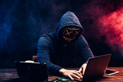 young criminal male hides his face under the hood and mask, hacks the password on the laptop, typing something. anonymous, incognito guy going to hack. cyberattack concept