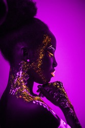 Portrait of african young woman with fluorescent makeup on face and skin that glowing in ultraviolet light. colorful, interesting and unusual shoot. side view. pink background