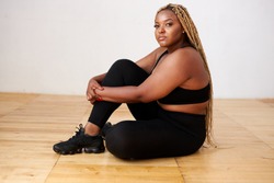 charming fat black female sit relaxing in studio with white background, beautiful woman in black sportswear look at camera, plus size model