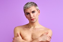 feminine shirtless man with make-up posing at camera, hugging himself, isolated on pink background