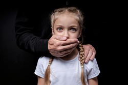 Scared girl with closed mouth by maniac killer, looking at camera with wide opened eyes, male hands on kid's shoulders, kidnapping, pedophile with child girl