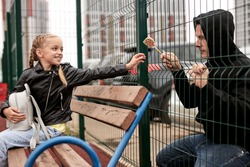 Stranger offers candy to child sitting on playground. Kid in danger. Children safety protection kidnapping concept. Smiling happy caucasian girl in leather jacket is affable, don't afraid of strangers