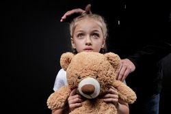 man kidnapping teenage girl. The concept of kidnapping and child trafficking. Close up portrait of frightened child in white t-shirt holding toy, looking at camera, isolated on black studio background