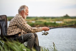 Senior man fishing outside in evening on lake in summer sitting on chair, enjoy spending time in nature, want to catch a fish, alone, side view portrait of male in casual checkered shirt