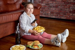Fat girl eating chips from bowl while sitting on floor in living room, side view. Overweight caucasian child in casual clothes enjoy leading unhealthy lifestyle, eat junk food and watch tv.