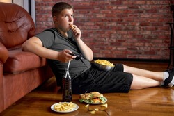 Fat teenager boy sitting on floor in living room, side view. Overweight obese caucasian child in casual clothes enjoy leading unhealthy lifestyle, eat junk food and play video games.