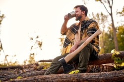 Young exhausted male sit drinking tea after wood cutting, lamberjack or lumberman, wood worker in casual clothes looking at side, having rest, taking a break at summer day alone. ecology, forestry