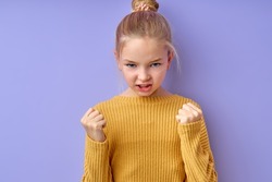 emotional girl in yellow shirt over isolated purple background Punching fist to fight, aggressive and angry attack, threat and violence. blonde kid girl is mad raising fist frustrated and furious