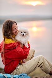 Woman with white spitz dog sit on top of mountain enjoying time during travel, beautiful pretty caucasian lady in sportswear at sunset in the evening. tourism, travel, adventure concept