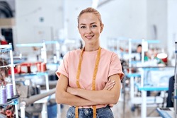positive woman dressmaker standing in textile factory at workshop, attractive tailor after work stand posing, looking at camera smiling. female use measuring tape for sewing and making garments